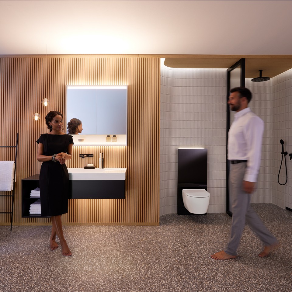 Un uomo e una donna in un bagno armoniosamente illuminato con mobili da bagno e ceramiche Geberit ONE (© Geberit)